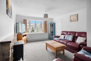 Ein Wohnzimmer mit einem roten Ledersofa, einem Holztisch, einer blauen Kommode und einem großen Fenster mit Vorhängen.