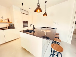Una cocina moderna con armarios blancos, encimera negra, horno empotrado y cafetera. Hay tres taburetes en la isla.