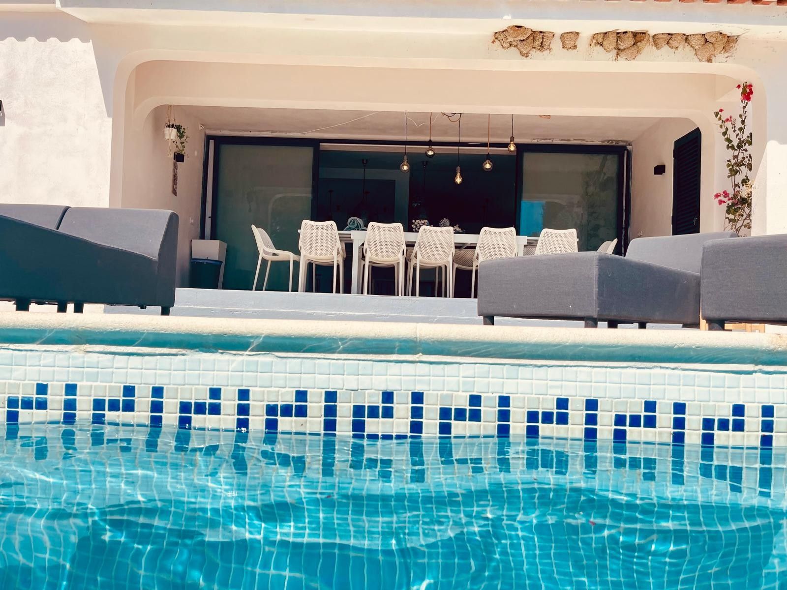 Un área de piscina con borde de azulejos, una mesa de comedor con sillas y una zona de estar cubierta.