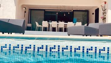 Un área de piscina con borde de azulejos, una mesa de comedor con sillas y una zona de estar cubierta.