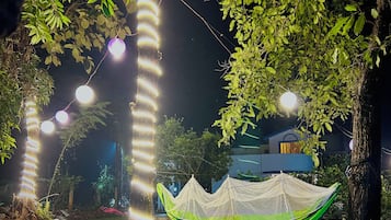 Un hamac suspendu entre deux arbres, un bâtiment en arrière-plan et des guirlandes lumineuses.