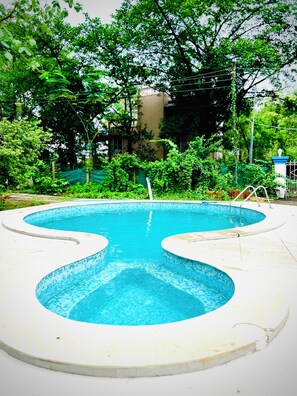 A swimming pool with a unique wavy edge design, surrounded by lush greenery and a fence. - Casa Santorini by Roamvacay (Kashele)