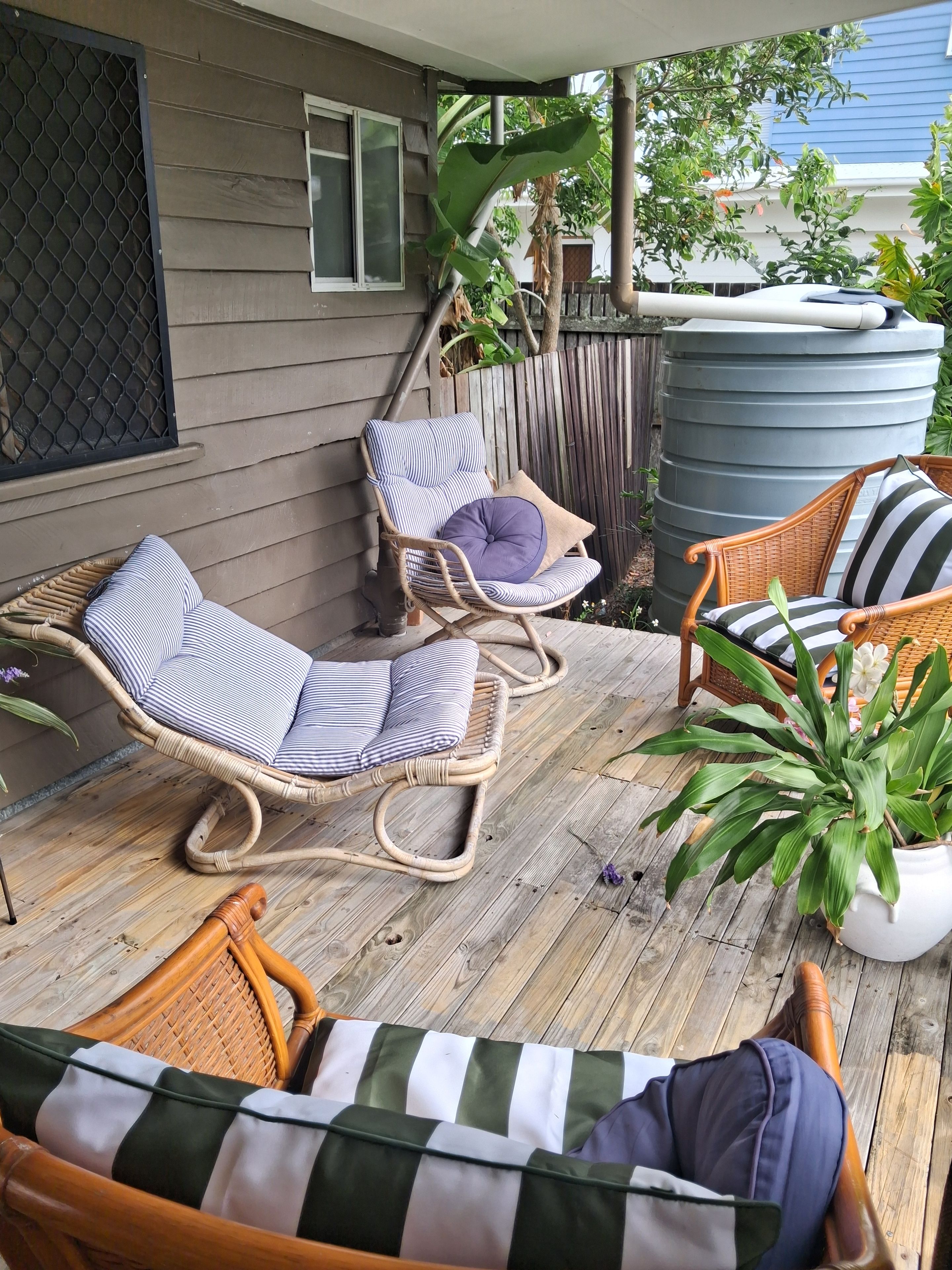 Una terraza de madera con mobiliario de mimbre, que incluye dos sillas y un sofá, y una planta grande en maceta.