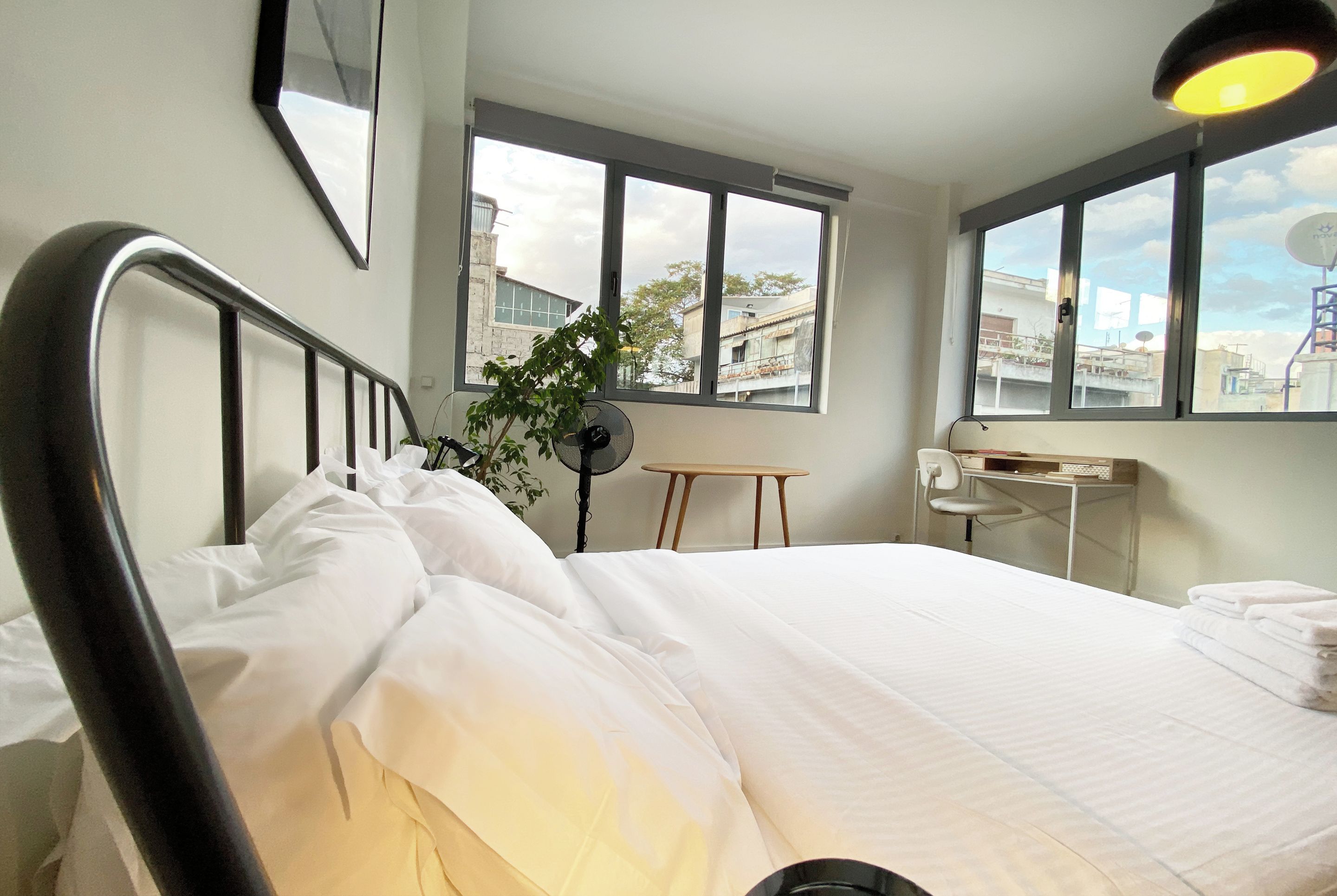 Hotellihuone, jossa on sänky, työpöytä, tuoli, viherkasvi ja suuret ikkunat.