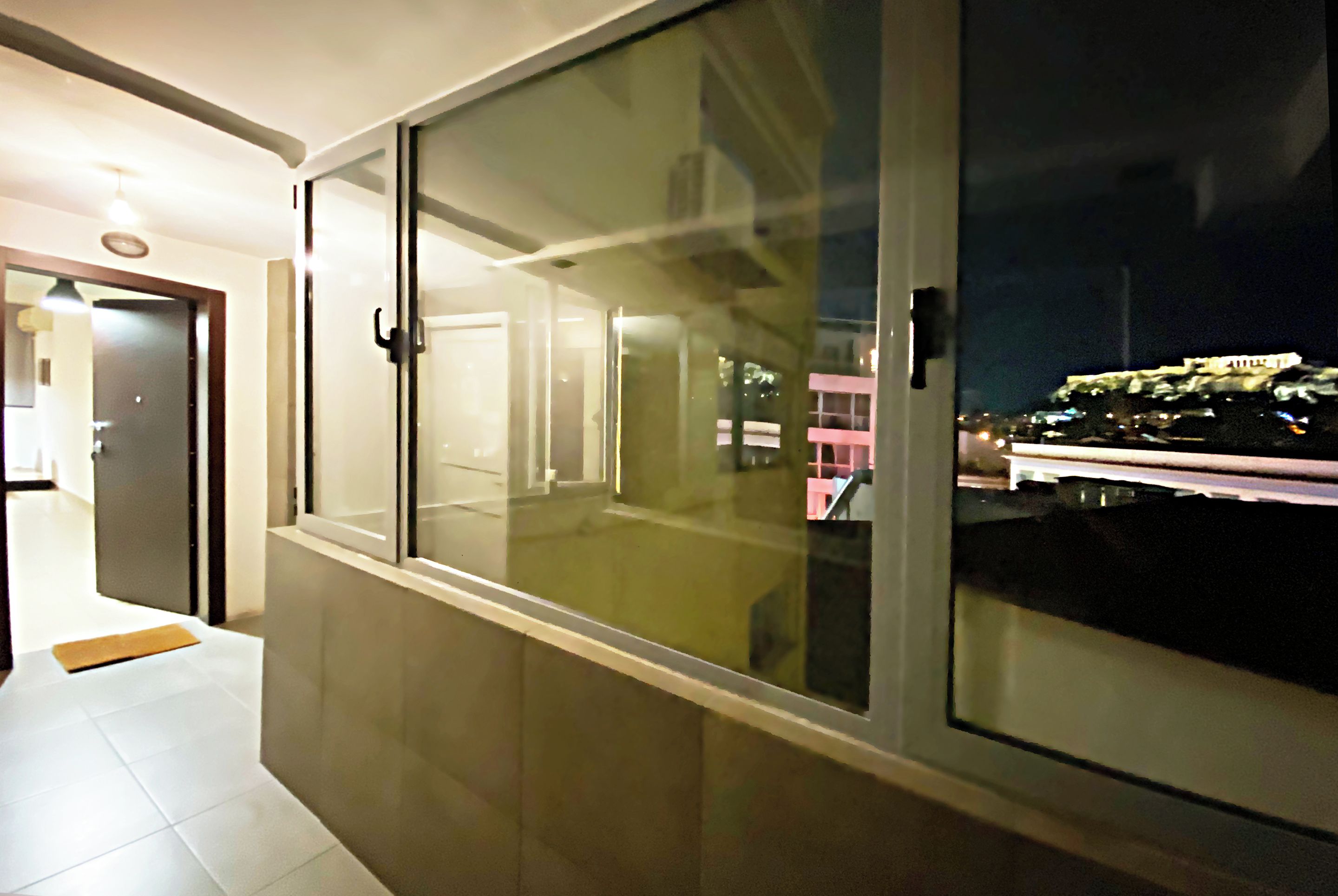 A corridor with a glass door leading to a balcony with a night view of a cityscape.