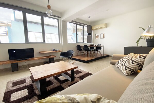 A modern living room with a sofa, a coffee table, a TV, and a dining area with chairs and a table. - Live in Athens Psyrri Lively Apartments (Athina)