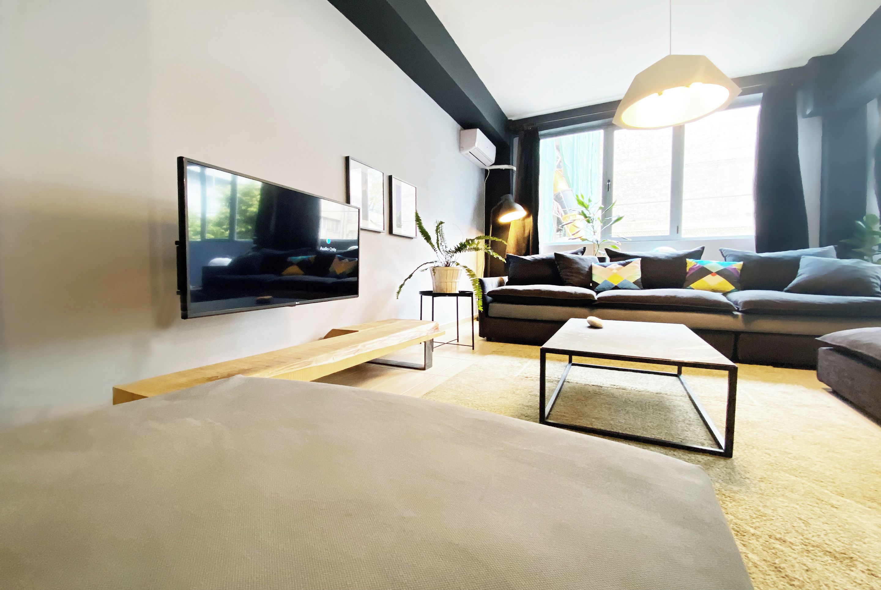 A modern living room with a flat-screen TV, a sofa, a coffee table, and a large window providing natural light.