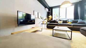 A modern living room with a flat-screen TV, a sofa, a coffee table, and a large window providing natural light.