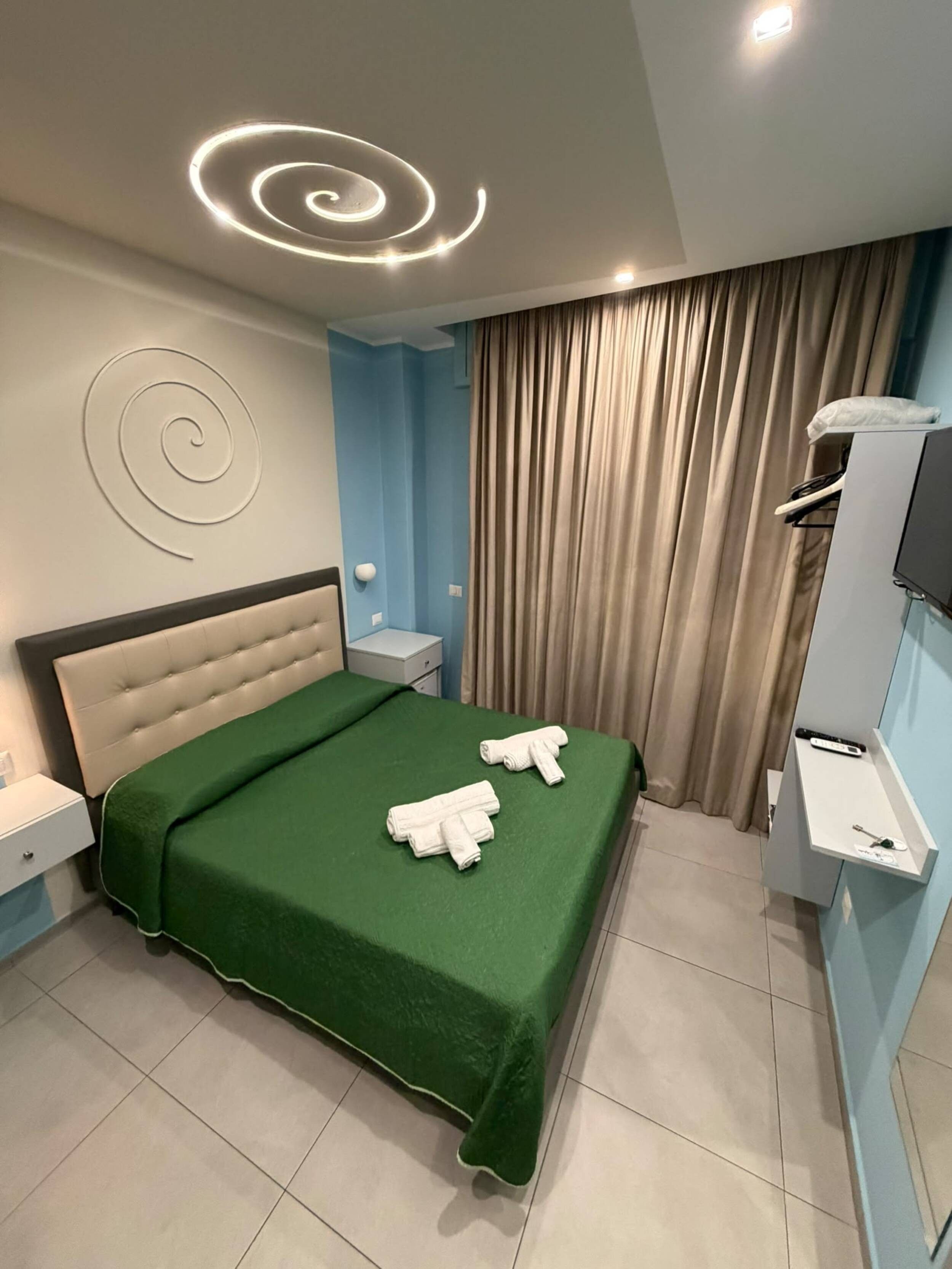 Hotelkamer met een bed, groene spreideken, twee handdoeken, een hoofdeinde, een nachtkastje, een televisietoestel en een gordijn.