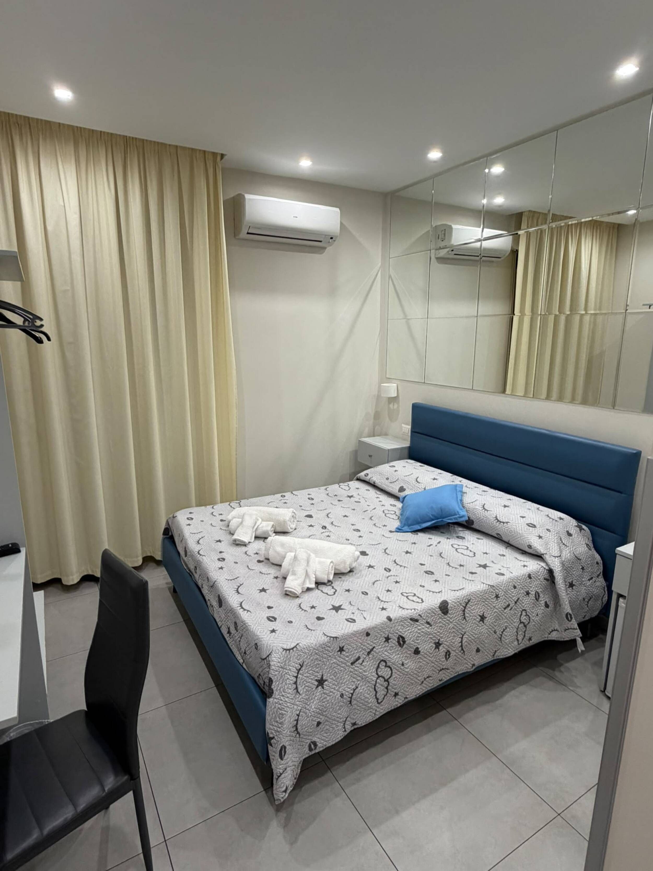 A hotel room with a bed, a desk, a chair, an air conditioning unit, and a mirror.