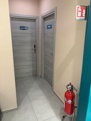 En hall med två dörrar, var och en märkt med namnen ”AFRODITE” och ”ATHENA”, en brandsläckare på höger sida och en skylt med texten ”ESTINTORE” ovanför.