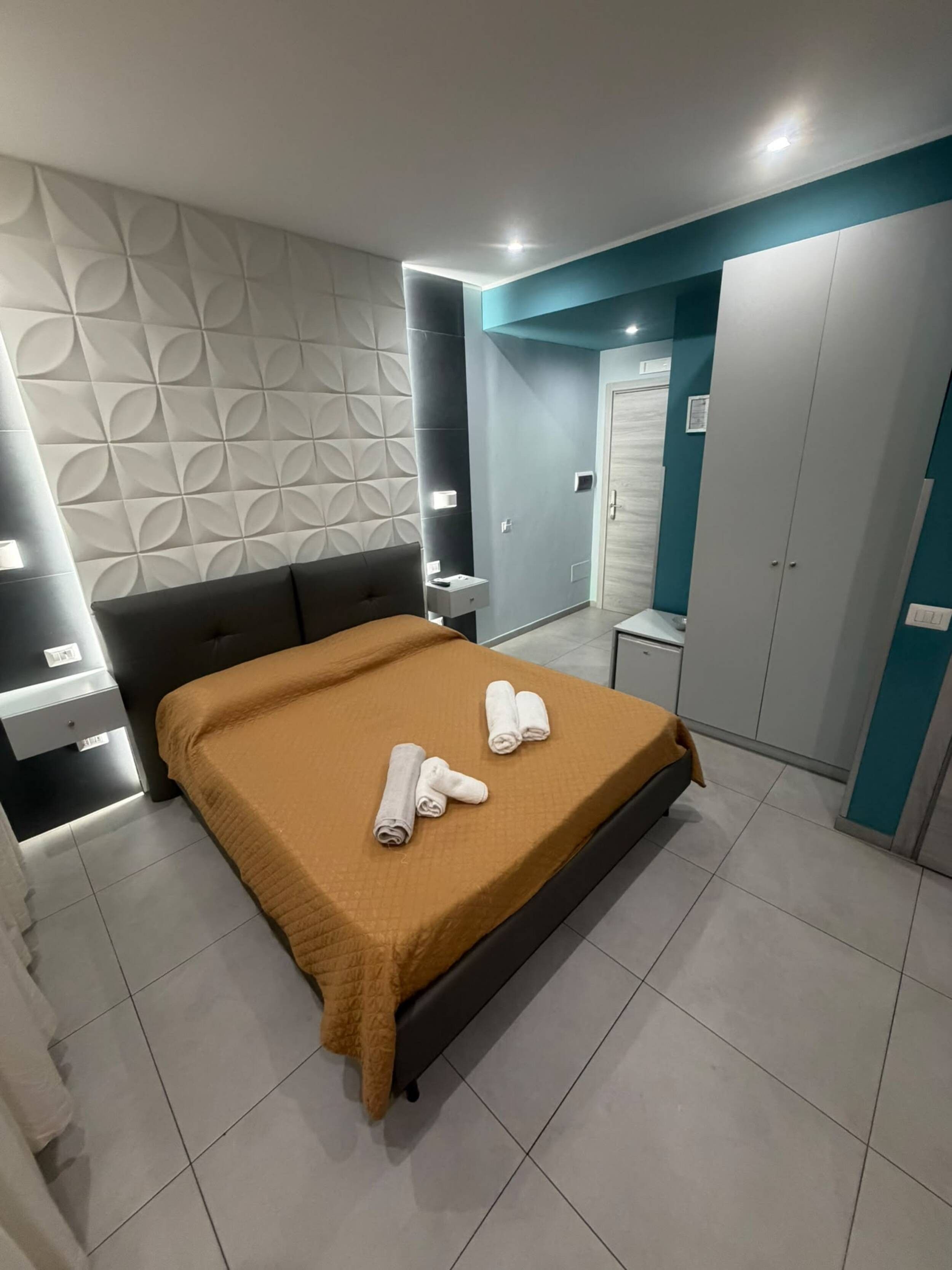 Une chambre à coucher moderne avec un lit, deux serviettes enroulées, une table de chevet, une armoire et une salle de bain visible au fond.