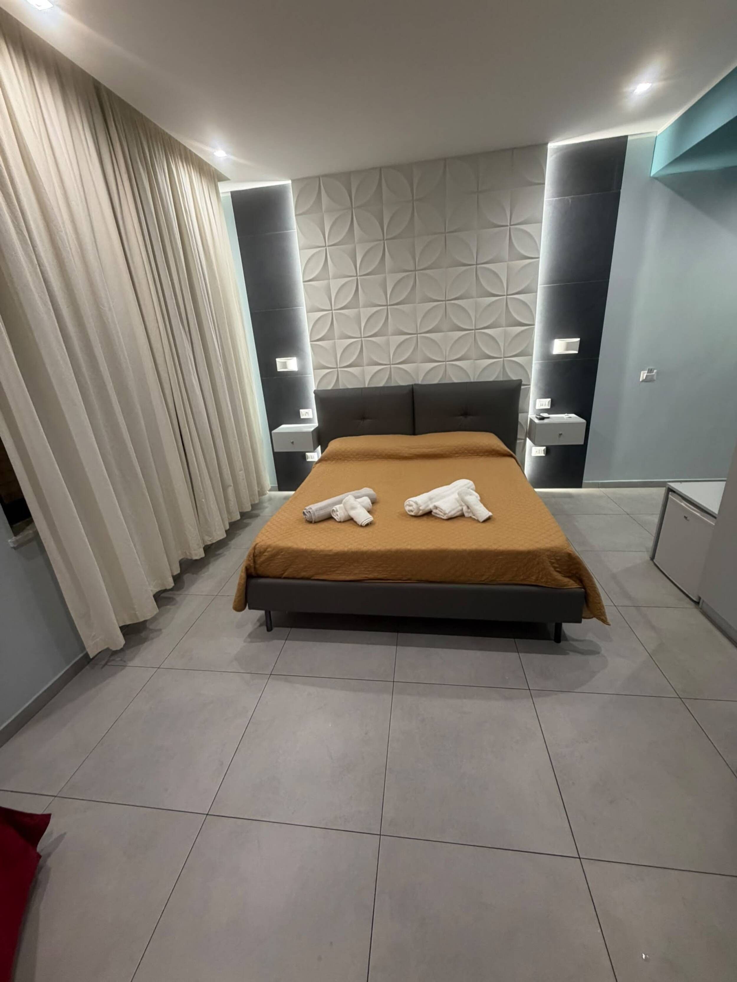A modern hotel room with a large bed, a headboard with a geometric pattern, and a wall-mounted nightstand.