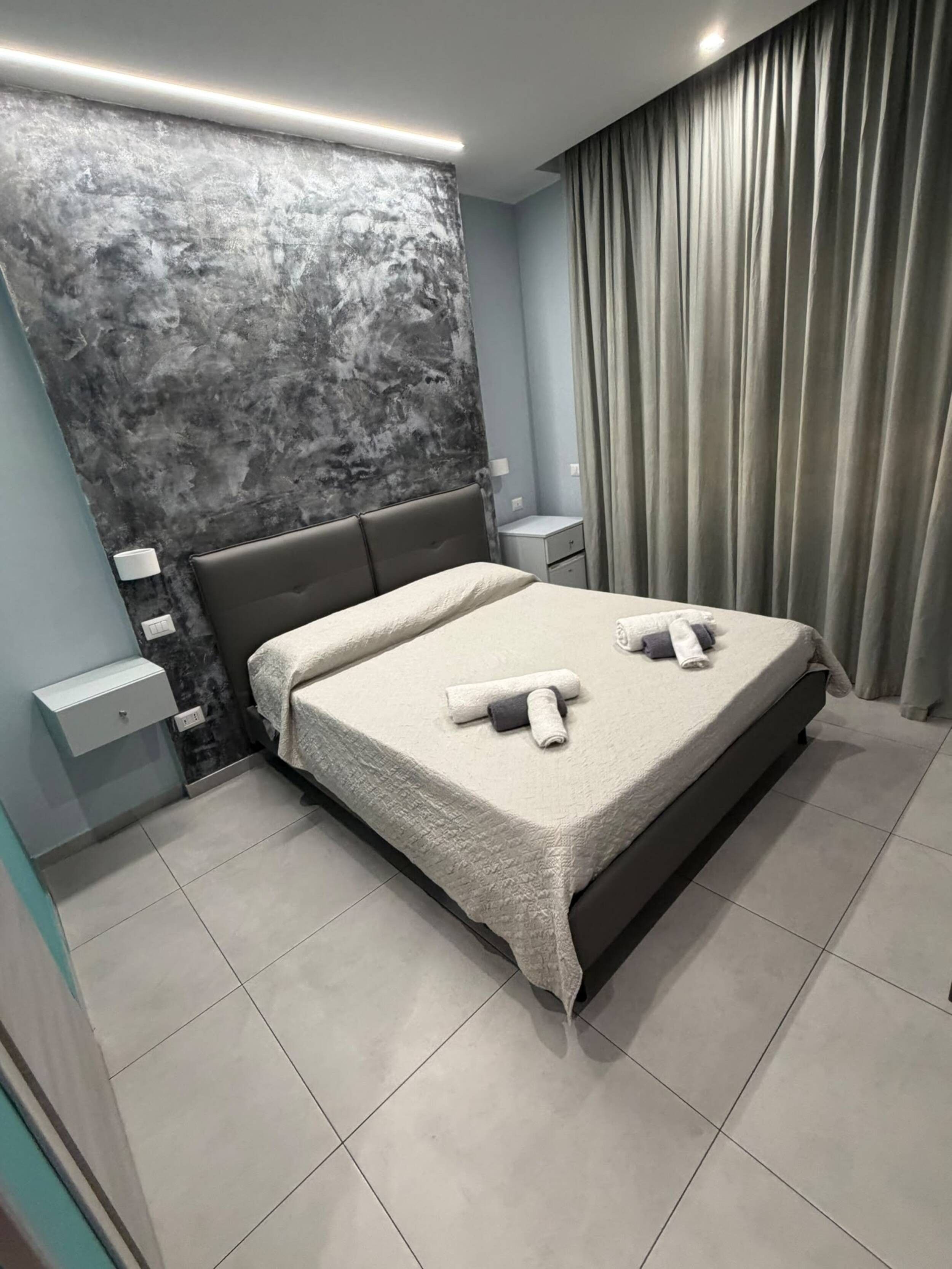 Un dormitorio moderno con una cama grande, una mesita de noche, una lámpara de pared y una cortina.
