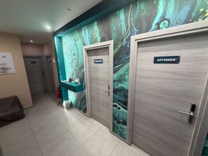 En hall med två dörrar märkta med namnen ”PERSEPHONE” och ”ARTEMIDE”, en turkosfärgad vägg med ett marmorerat mönster och en liten turkos hylla med diverse föremål.