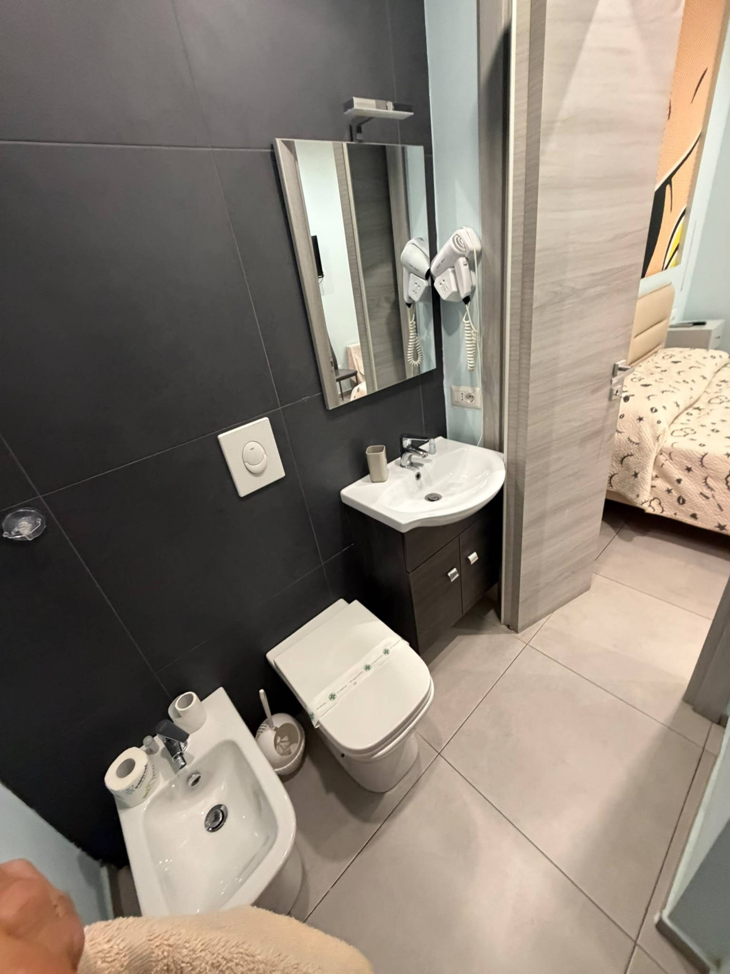 Un bagno moderno con una parete rivestita in piastrelle scure, un lavandino bianco, un bidet, uno specchio e un asciugacapelli.