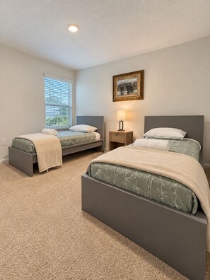 Quarto com duas camas de solteiro, mesa de cabeceira, abajur e janela com persianas.