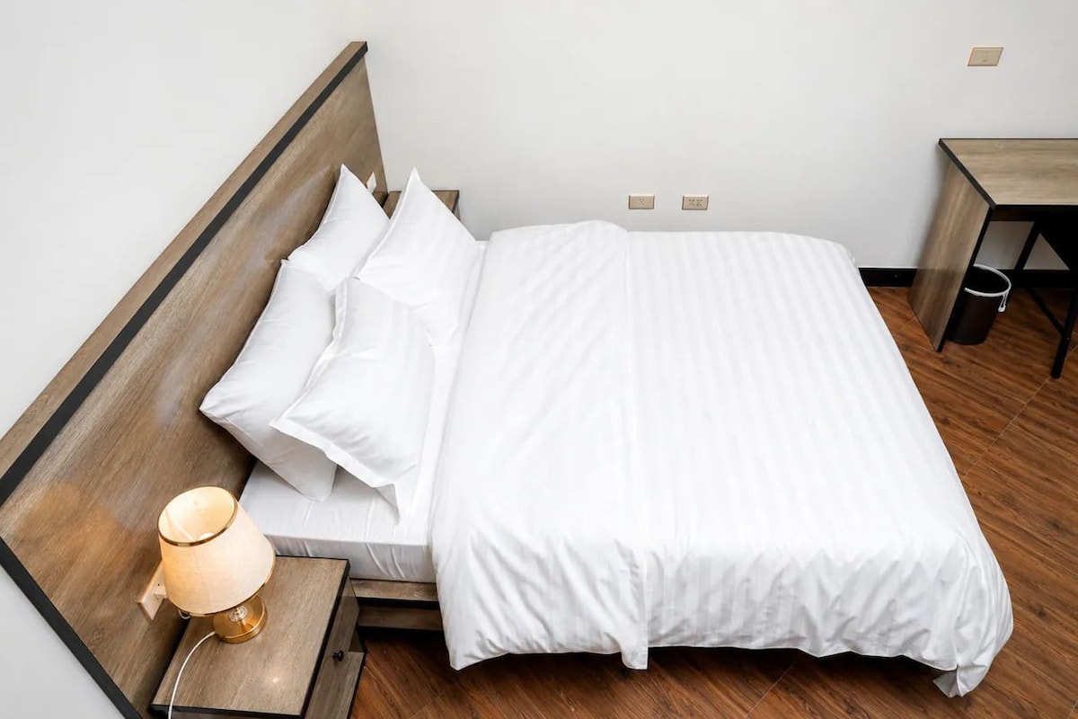 Un lit bien fait, avec du linge de lit blanc et des oreillers, une lampe de chevet et une table de nuit en bois.