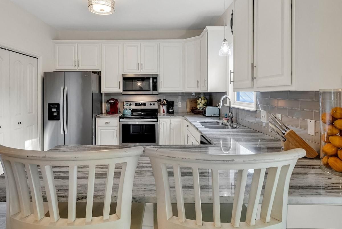 A modern kitchen with white cabinetry, stainless steel appliances, a marble countertop, and a dining area with two white chairs.