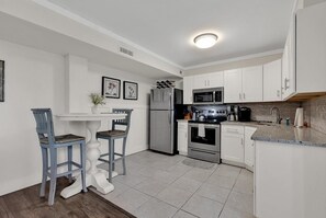 Eine moderne Küche mit weißen Schränken, einem Edelstahlkühlschrank, einer Mikrowelle, einem Backofen und einem Spülbecken. Zwei Barhocker und ein kleiner Tisch mit Stühlen sind ebenfalls vorhanden.