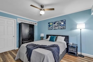 A bedroom with a bed, a ceiling fan, a wardrobe, artwork on the wall, and a bedside lamp.