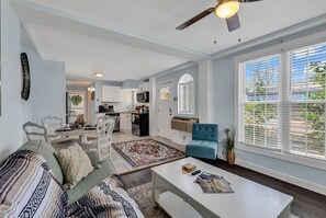 A modern living room with a sofa, coffee table, and a blue armchair. There is a kitchen in the background with white cabinets and a microwave. - Marshwalk Retreat! 2 Bedroom Hideaway! (Murrells Inlet)