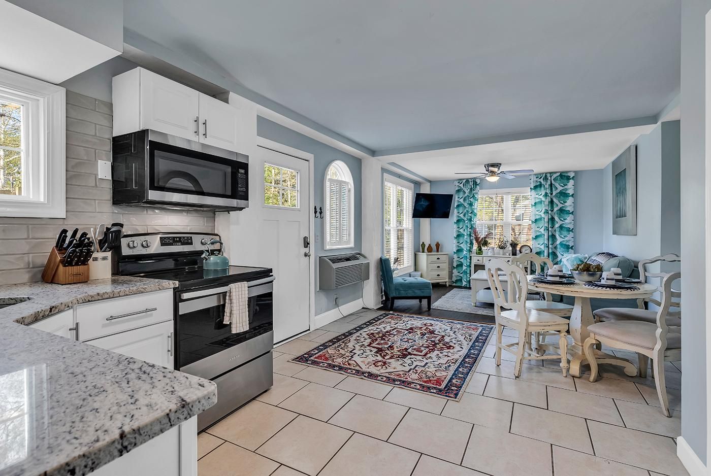 A modern kitchen with white cabinetry, a built-in microwave, and a stove. There is a dining area with a table and chairs, and a patterned rug on the floor.