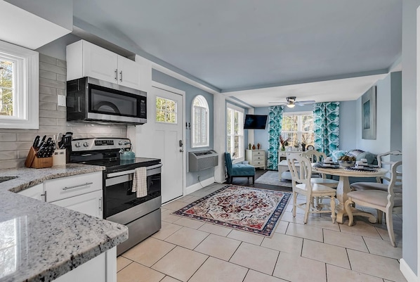 A modern kitchen with white cabinetry, a built-in microwave, and a stove. There is a dining area with a table and chairs, and a patterned rug on the floor. - Marshwalk Retreat! 2 Bedroom Hideaway! (Murrells Inlet)