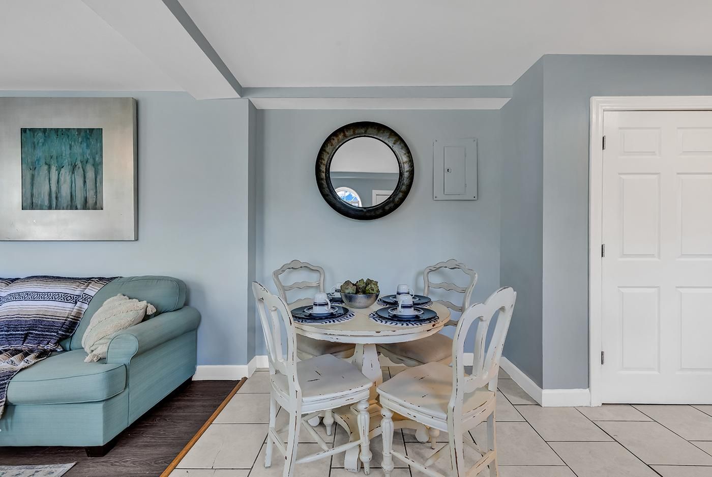 A dining area with a round table, white chairs, and a blue and white patterned tablecloth. There is a light blue sofa with a patterned throw blanket and a framed artwork on the wall.