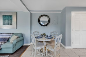 Zona pranzo con un tavolo rotondo, sedie bianche e una tovaglia a tema blu e bianco. Presente anche una poltrona blu chiaro con una coperta decorata e un quadro appeso al muro.
