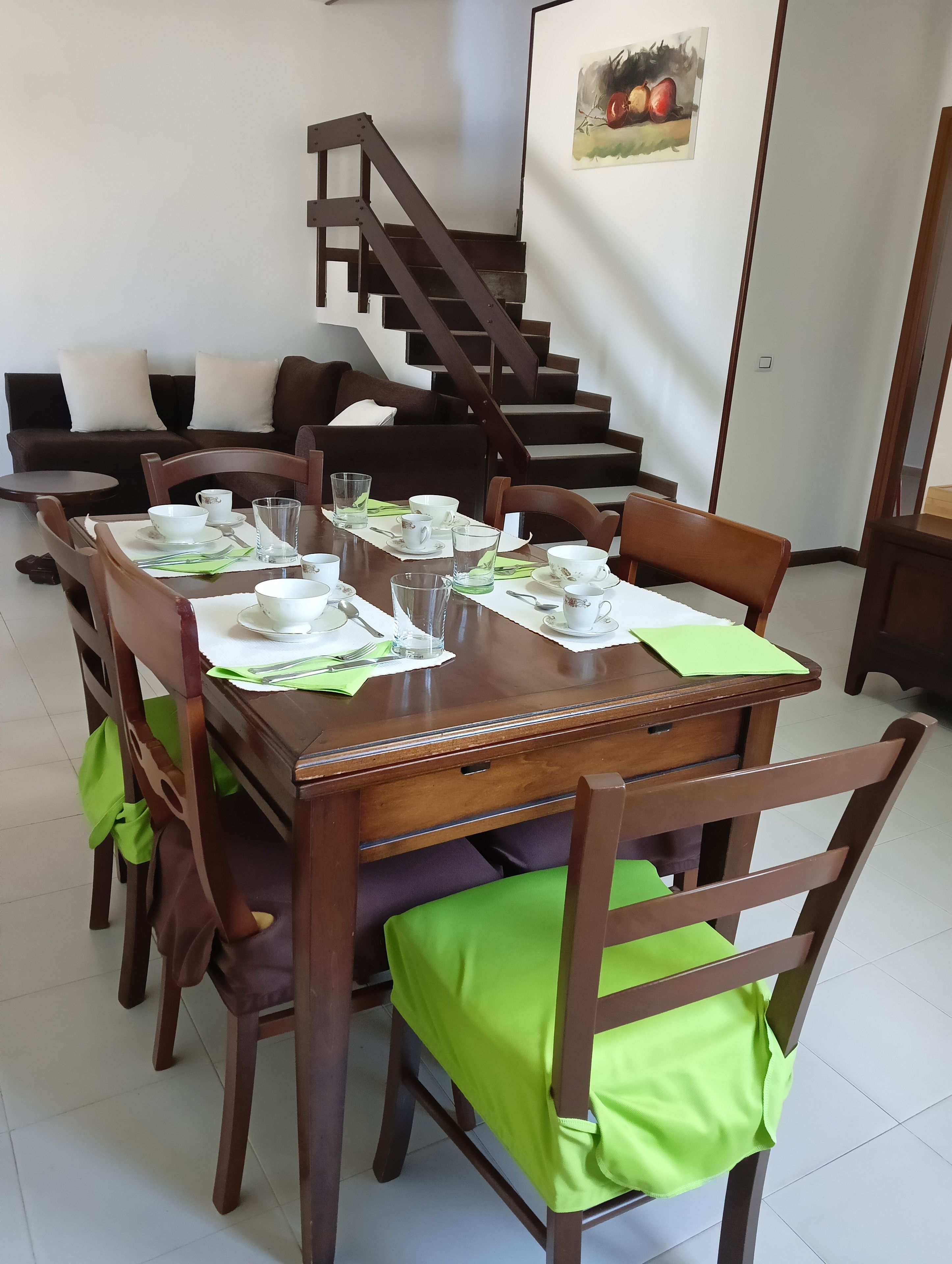 Un coin repas avec une table en bois dressée pour quatre personnes, entourée de chaises dotées de coussins verts. Un escalier est visible au fond.