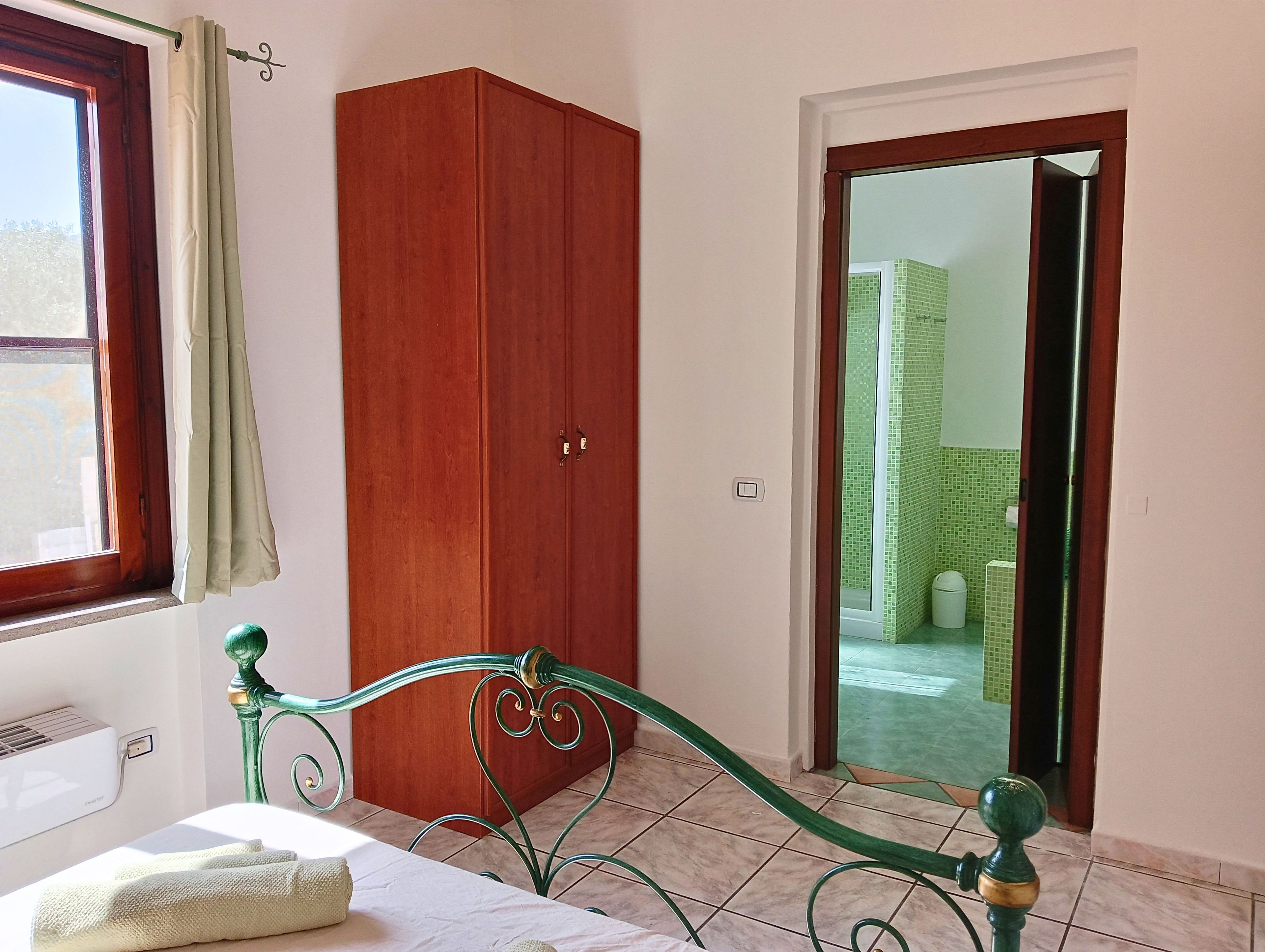 Quarto com cama, guarda-roupa, janela com cortinas e vista para uma casa de banho com azulejos através de uma porta aberta.