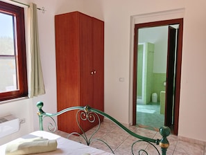 Ein Schlafzimmer mit einem Bett, einem Kleiderschrank, einem Fenster mit Vorhängen und Blick auf ein gefliestes Badezimmer durch eine offene Tür.