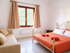 Un dormitori amb un llit, una tauleta de nit, una finestra amb cortines, un radiador i un sofà.