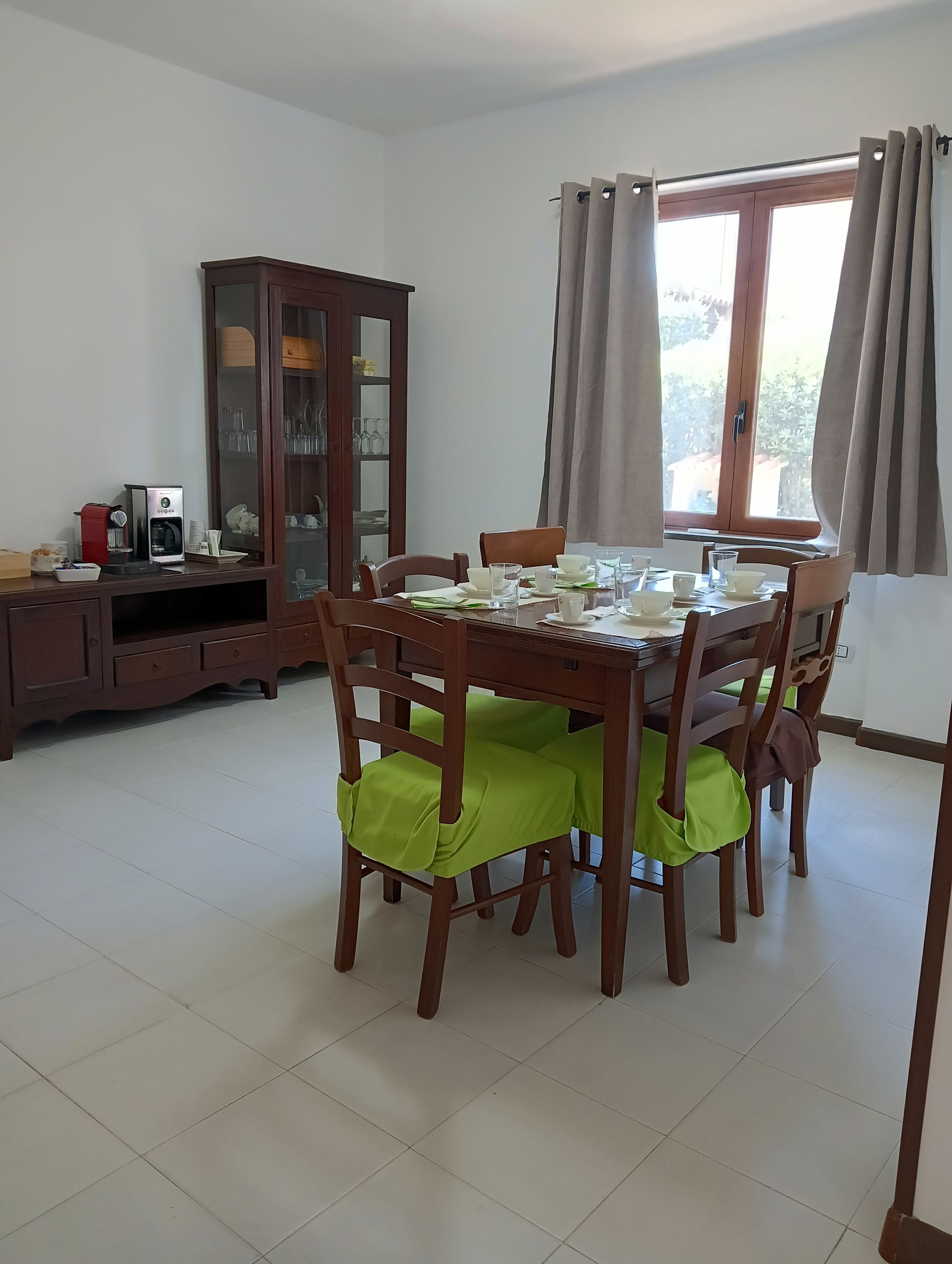 A dining area with a wooden table set for four, surrounded by chairs with green seat covers. A cabinet with glass doors is to the left, and a window with curtains is to the right.