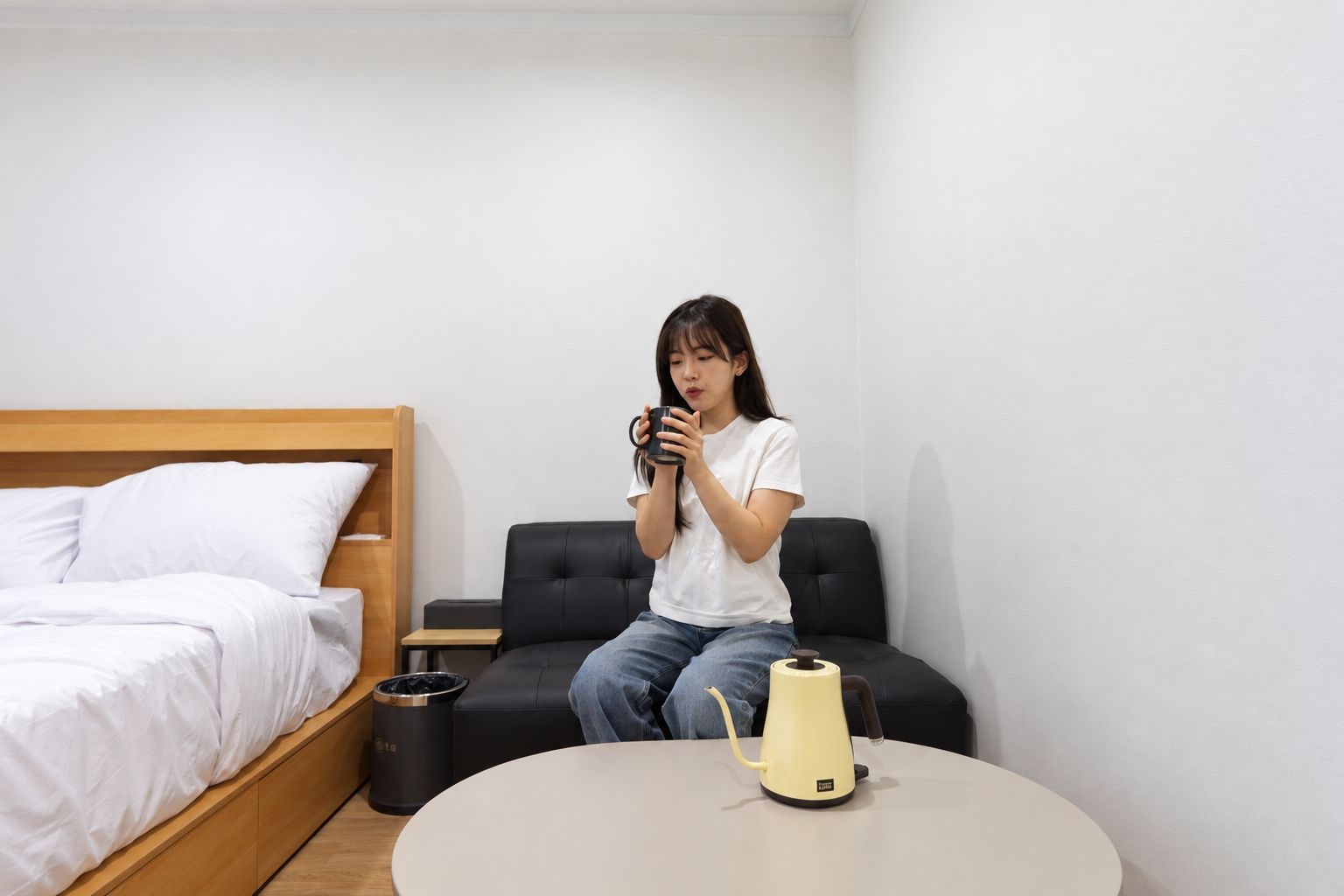 A room with a bed, a black sofa, a small table with a yellow kettle, and a trash bin.