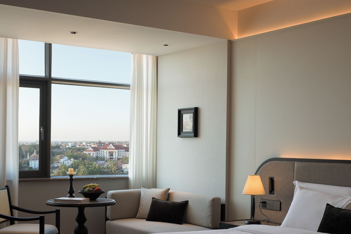 A hotel room with a large bed, a bedside table with a lamp, a small table with a book and a candle, a chair, and a view of the city through a large window.
