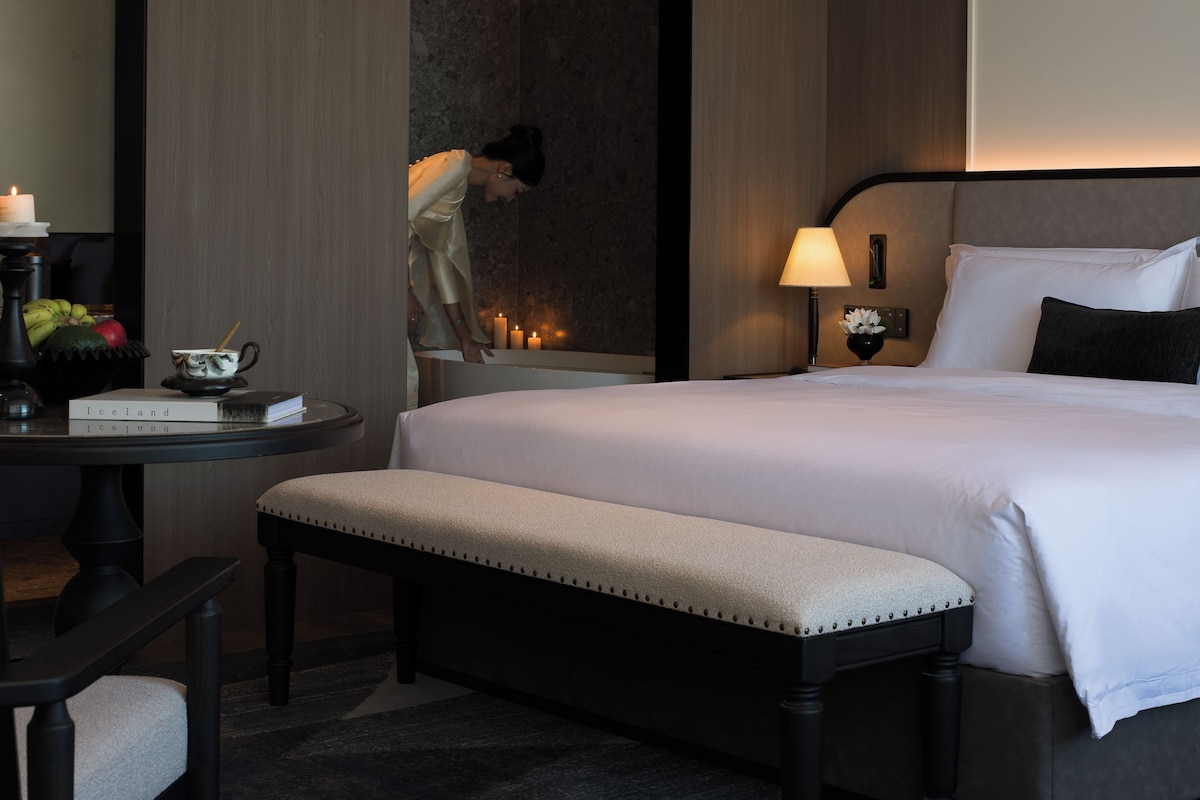 A hotel room with a large bed, bedside table, bench, and a mirror.