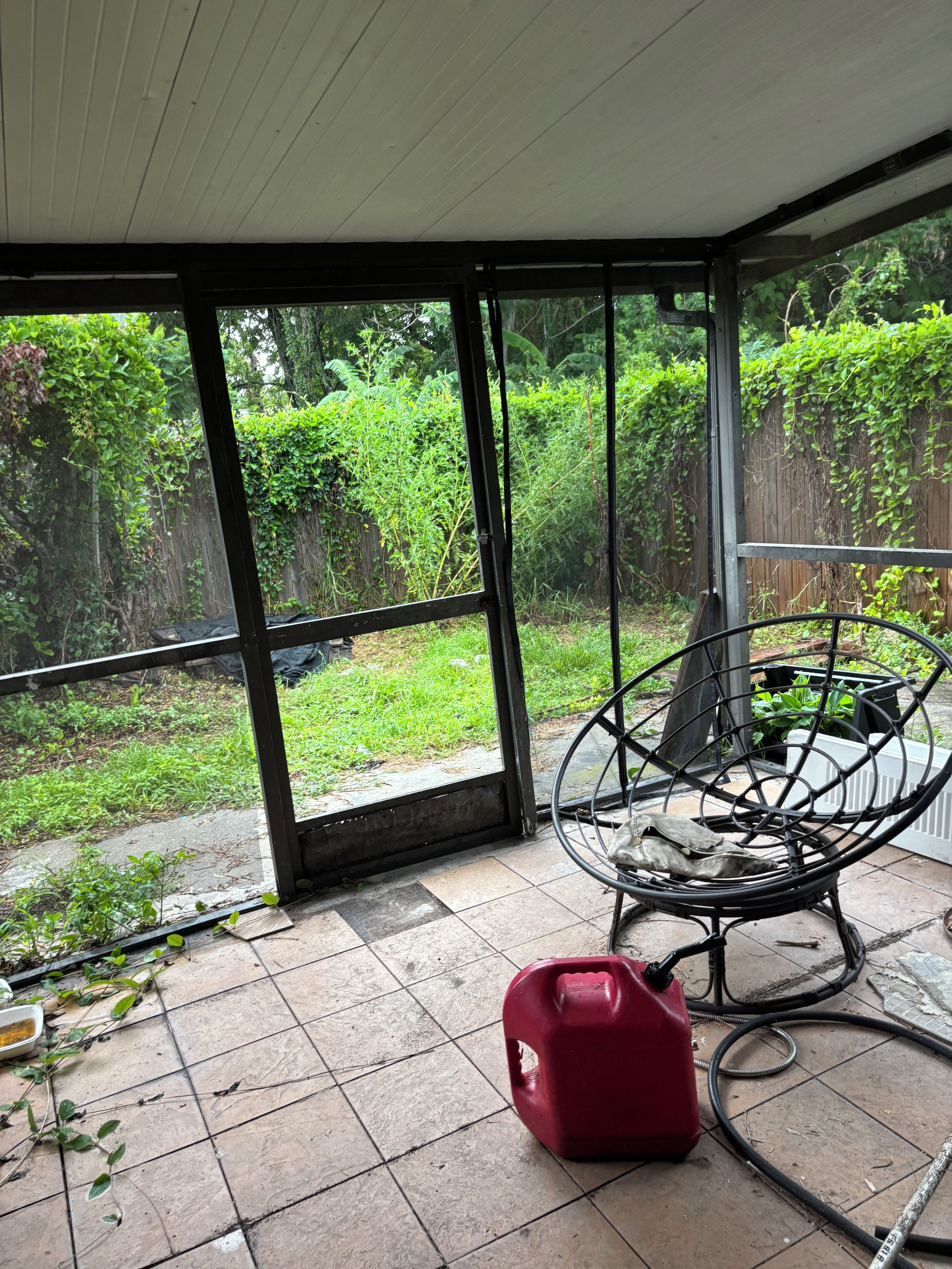 A room with a large glass door leading to a garden, a metal chair, and a red container on the floor.