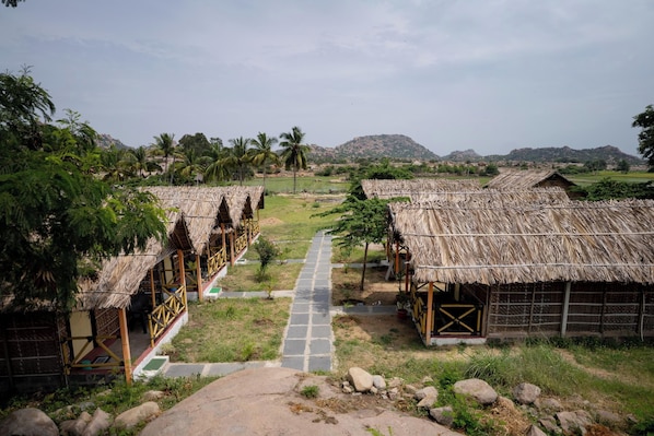 A row of thatched-roof huts with a paved pathway leading to them, set in a rural landscape with hills in the background.