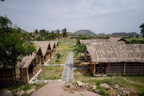A row of thatched-roof huts with a paved pathway leading to them, set in a rural landscape with hills in the background.