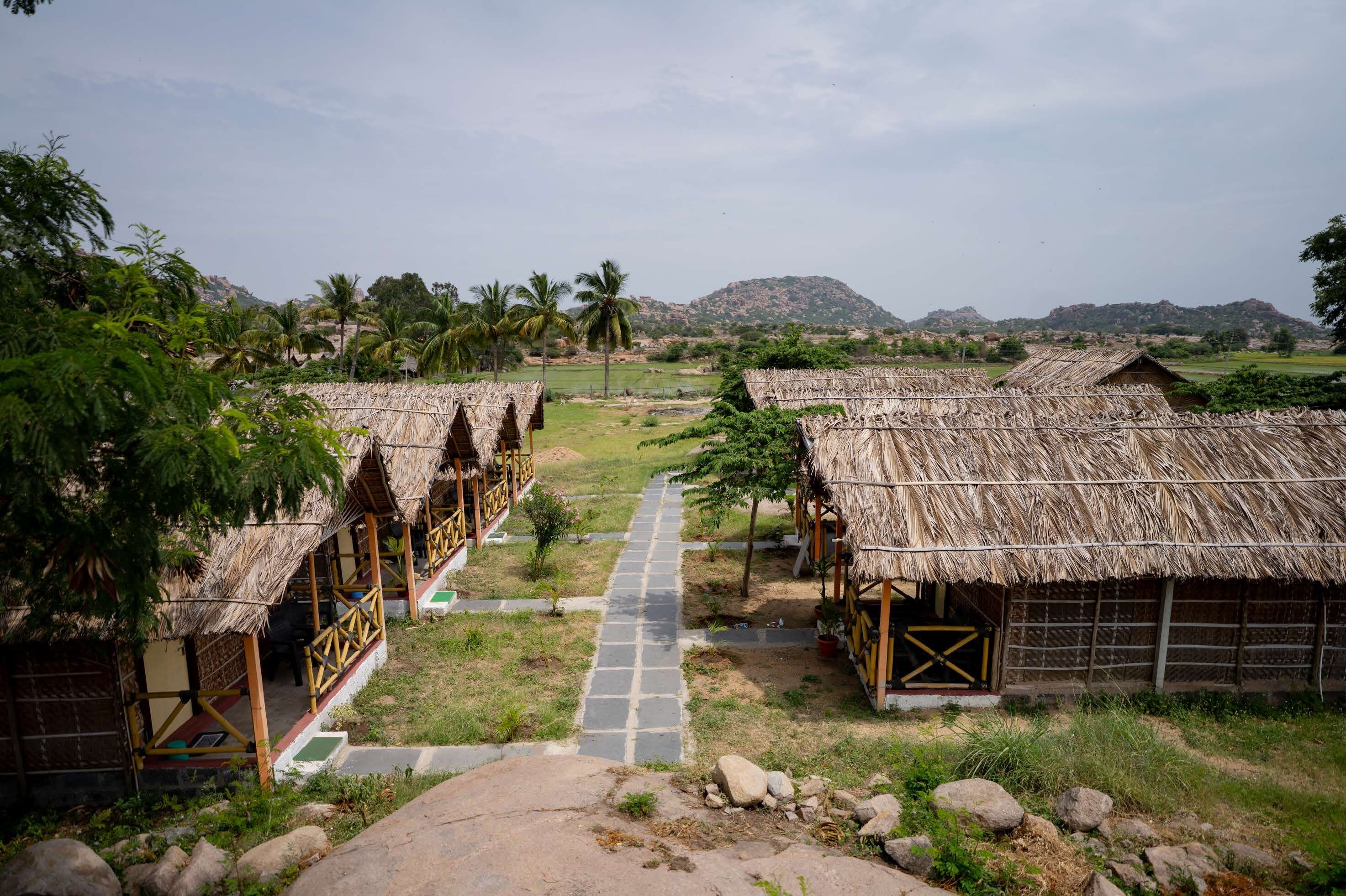 A row of thatched-roof huts with a paved pathway leading to them, set in a rural landscape with hills in the background.