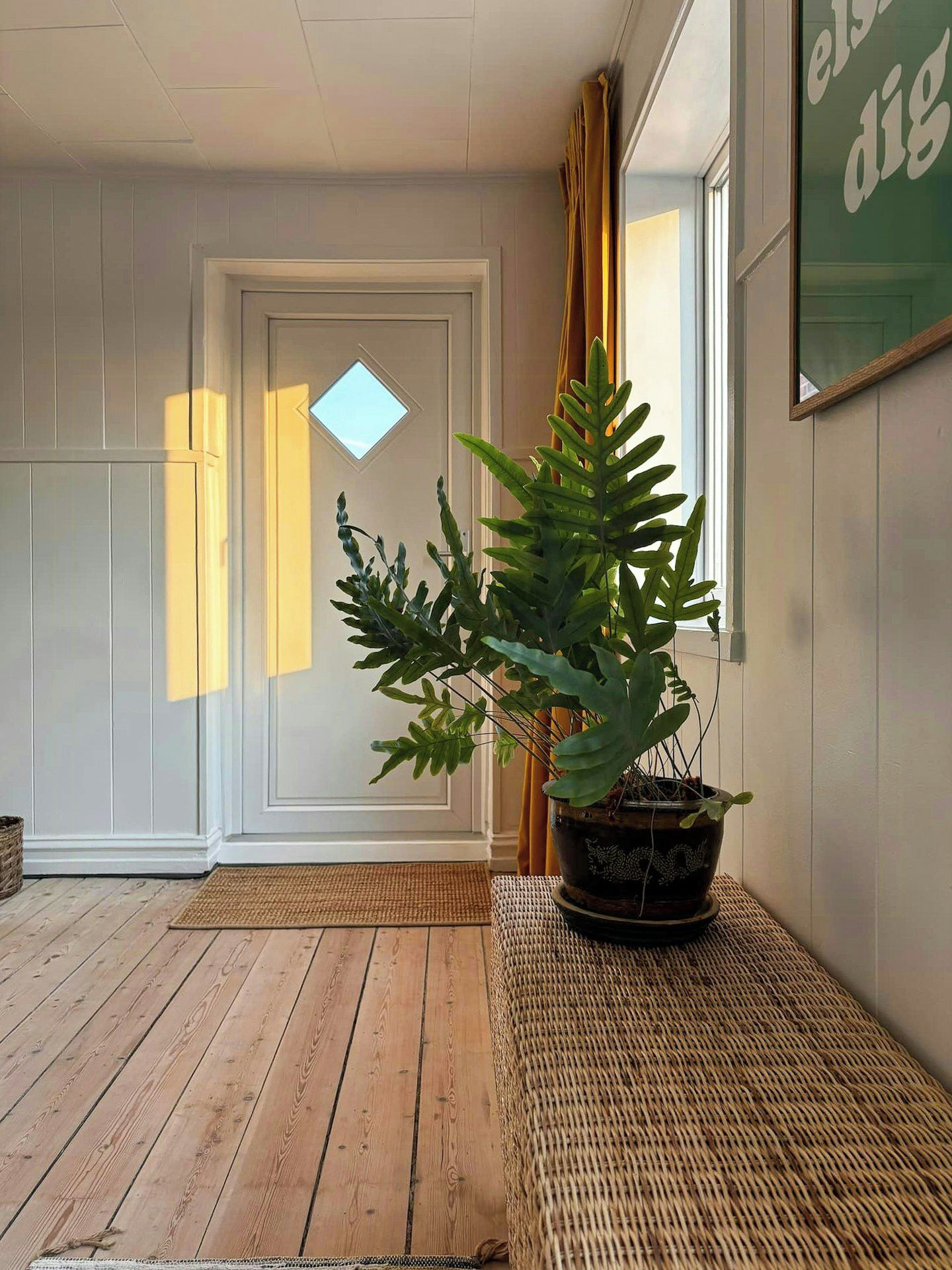 A room with a wooden floor, a potted plant, a door with a window, and a wall with a framed text.