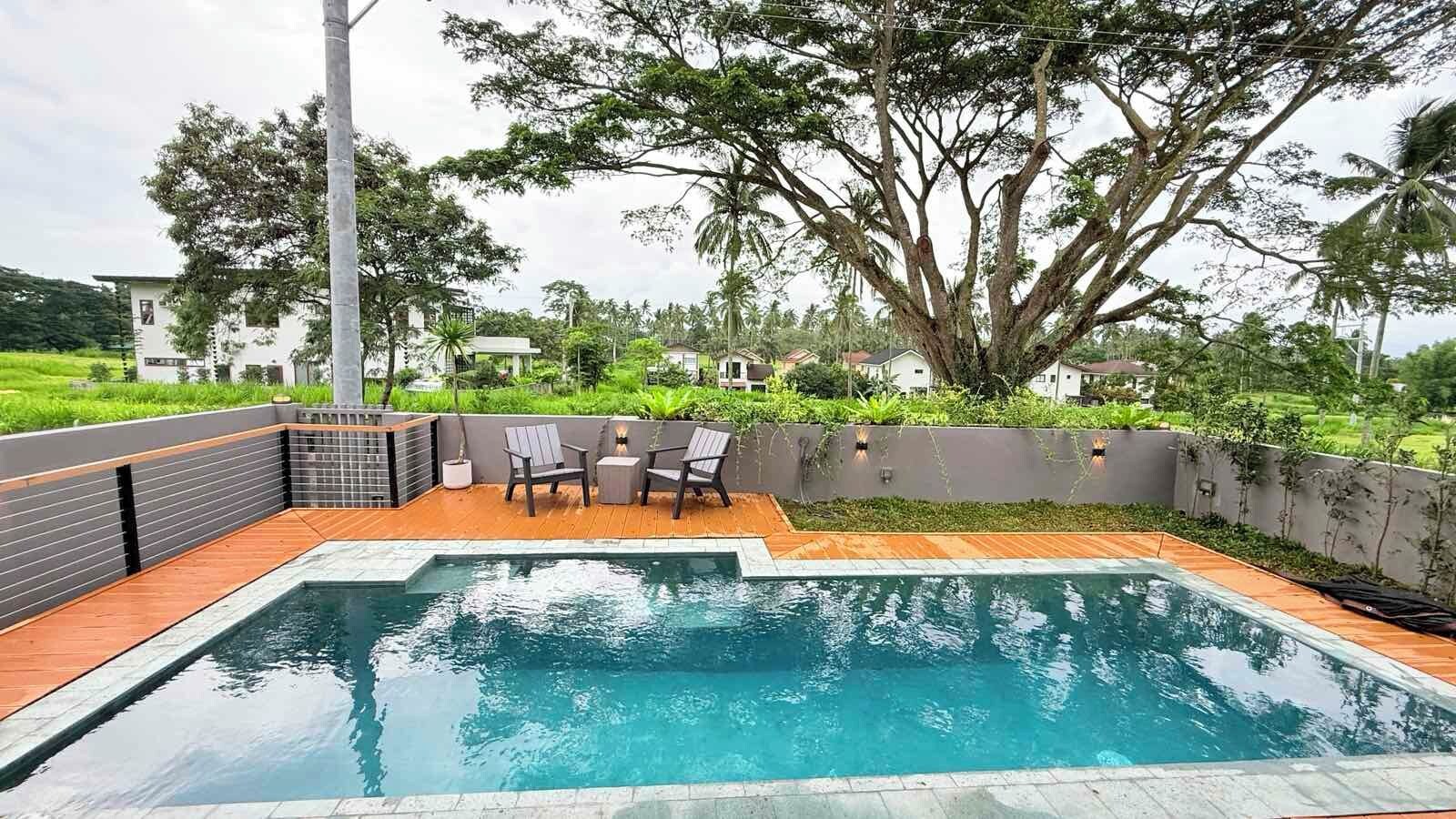 Piscina com deck de madeira e cadeiras, rodeada por vegetação e uma grande árvore.