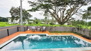Piscina com deck de madeira e cadeiras, rodeada por vegetação e uma grande árvore.