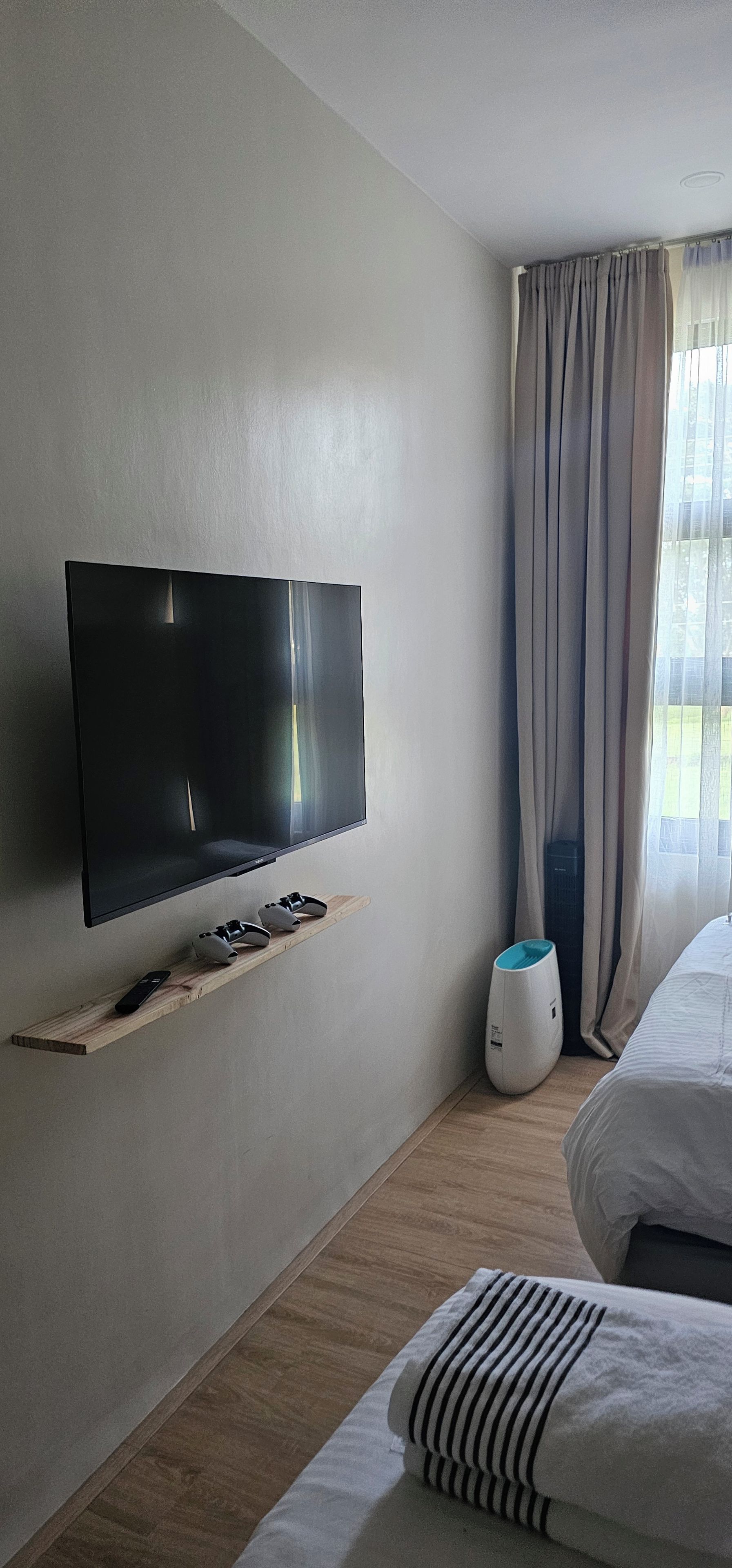 Quarto de hotel com televisão de tela plana na parede, prateleira de madeira com controle remoto, purificador de ar branco e cama com roupa de cama listrada.