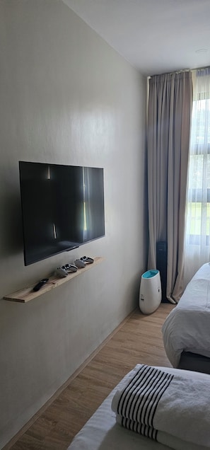 A hotel room with a flat-screen TV mounted on the wall, a wooden shelf with a remote control, a white air purifier, and a bed with striped bedding. - Avodah house at summit point golf and country club (Lipa City)