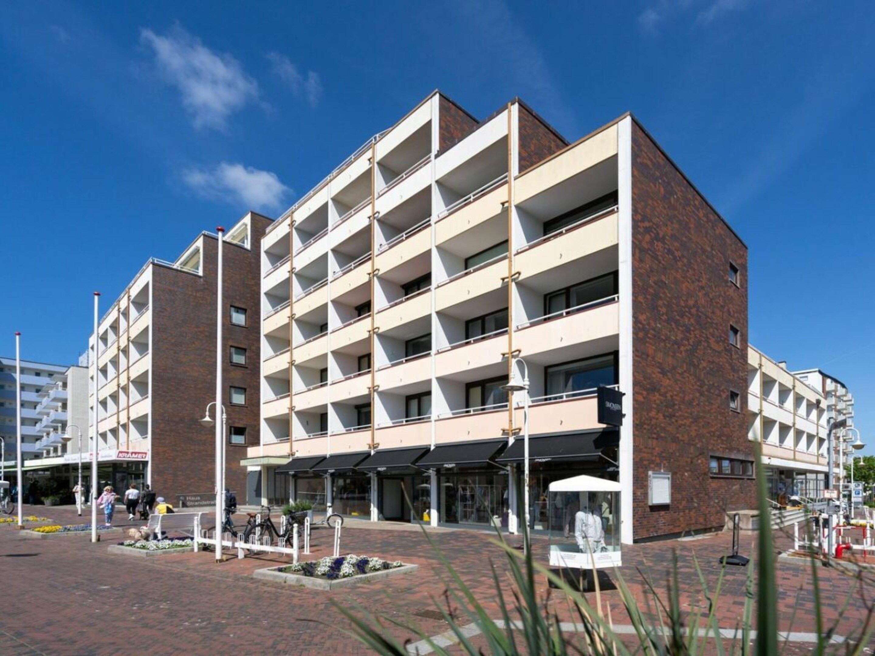 A multi-story building with a brick facade, multiple balconies, and a paved courtyard with benches.
