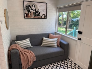 A grey sofa with striped pillows and a brown throw blanket, a framed picture on the wall, and a window with a view of greenery.