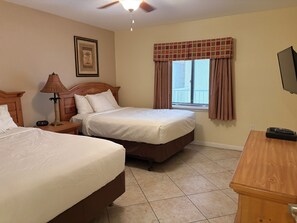 Habitación de hotel con dos camas, ventilador de techo, ventana con cortinas, un cuadro en la pared y un televisor sobre un soporte de madera.